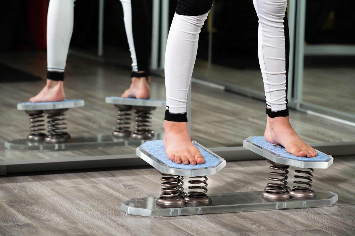 A dancer's feet in relevé on a double spring balance board with blue tops in a mirrored studio