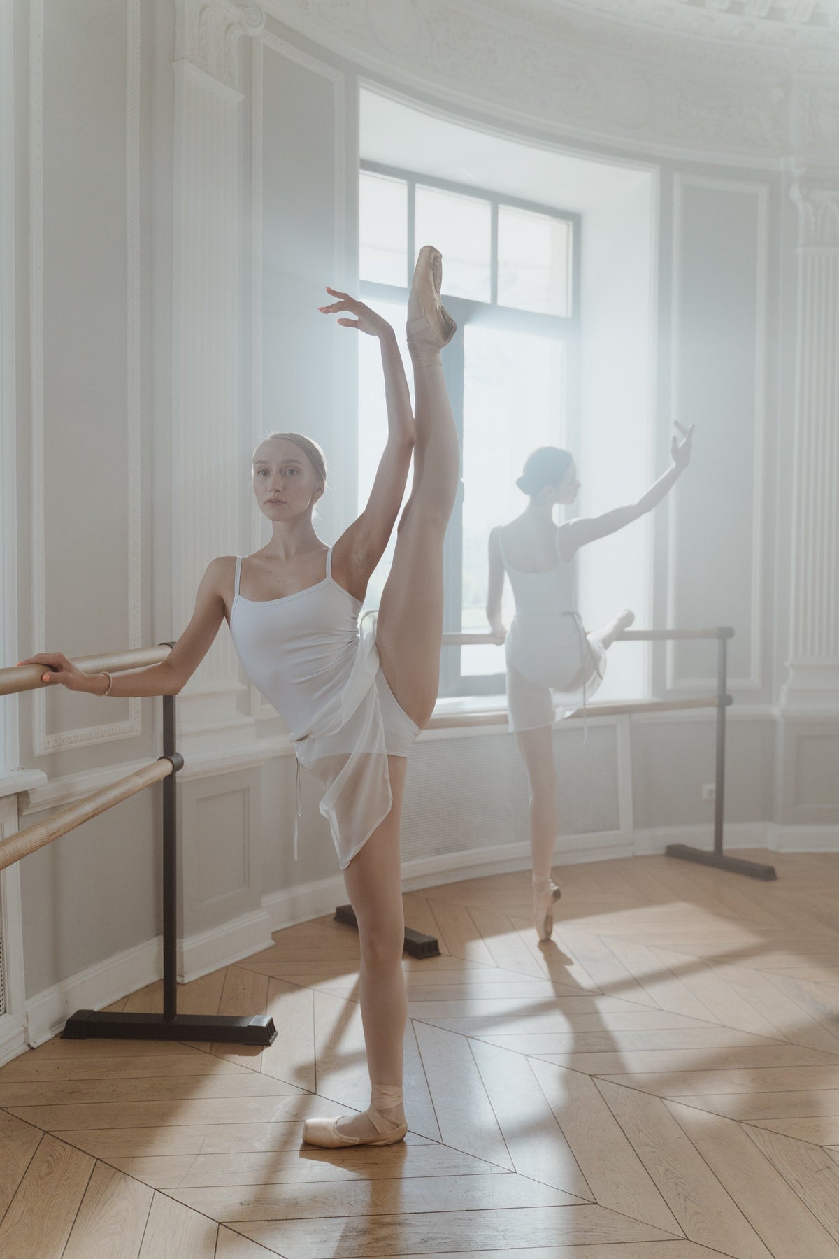 A ballet dancer in a white leotard holding a high développé at the barre in a bright studio, with a second dancer behind her