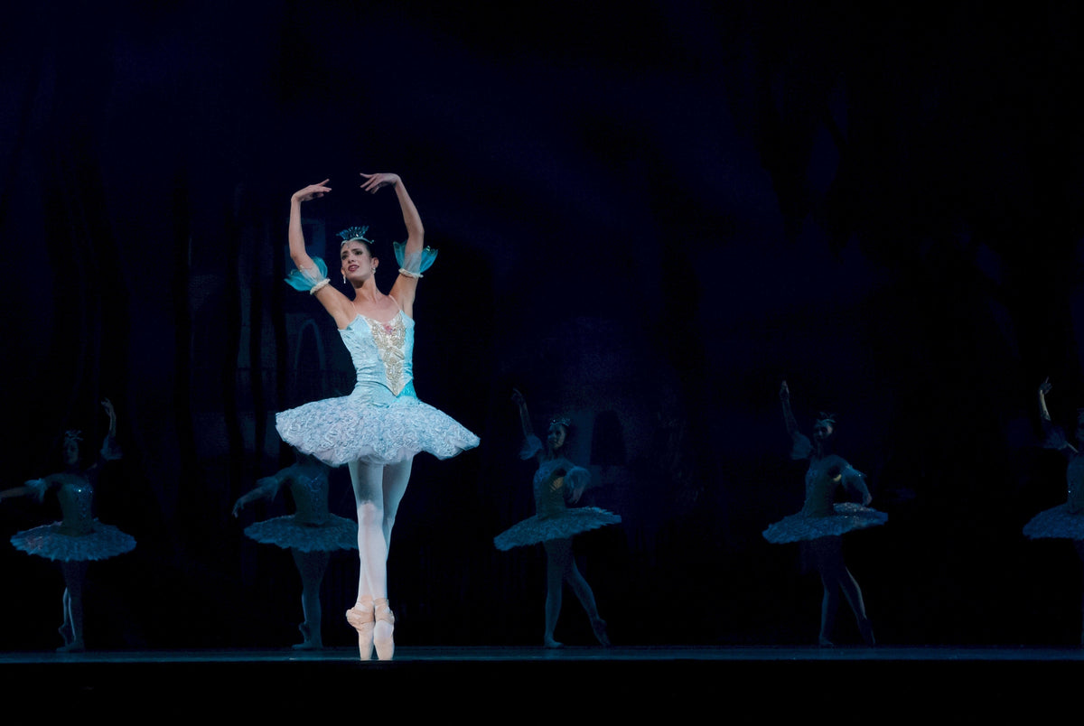 Ballerina in a white tutu dancing on a dark stage with the corps de ballet behind her