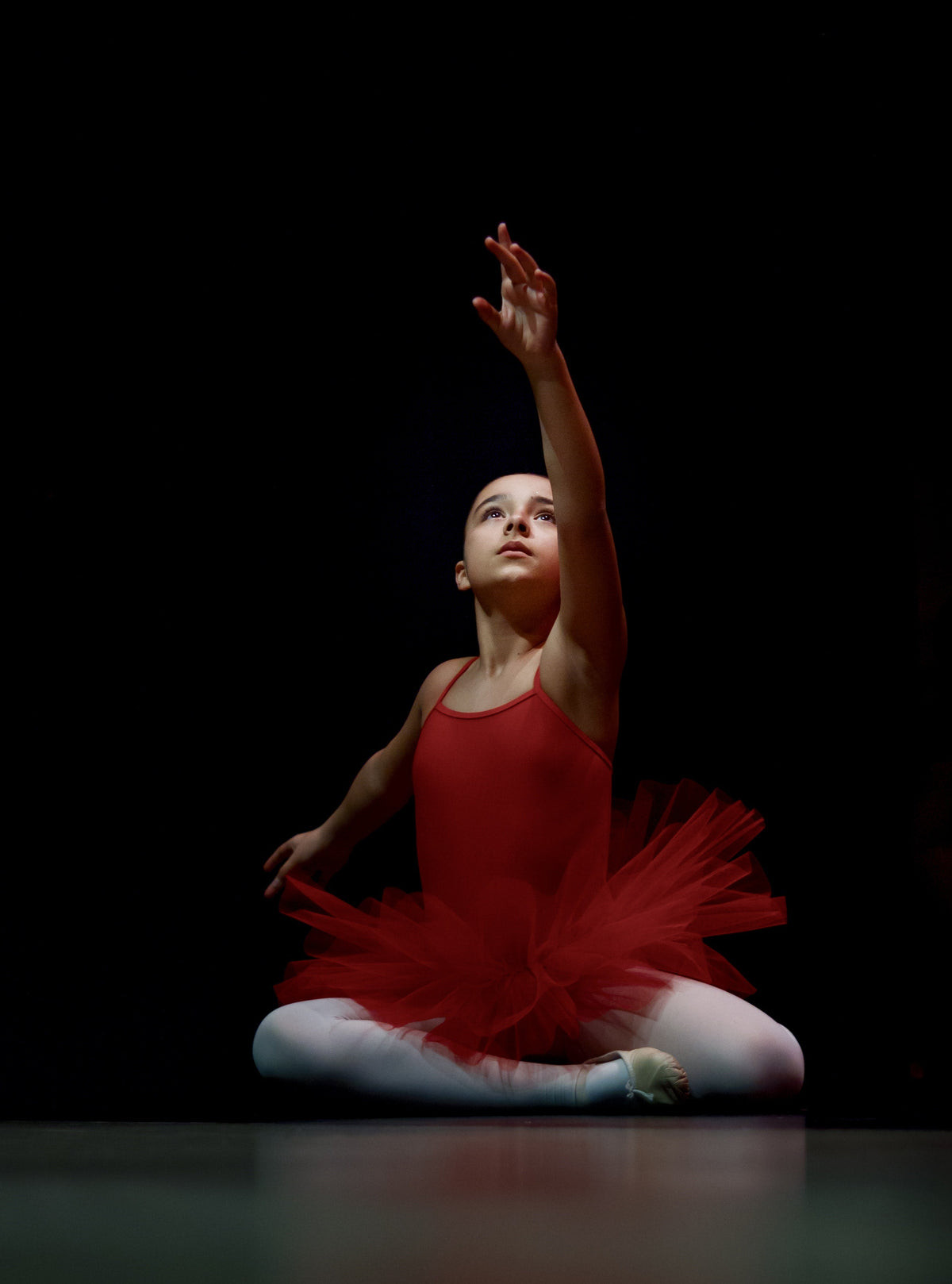 Young girl in a red tutu seated on a dark stage with one arm raised